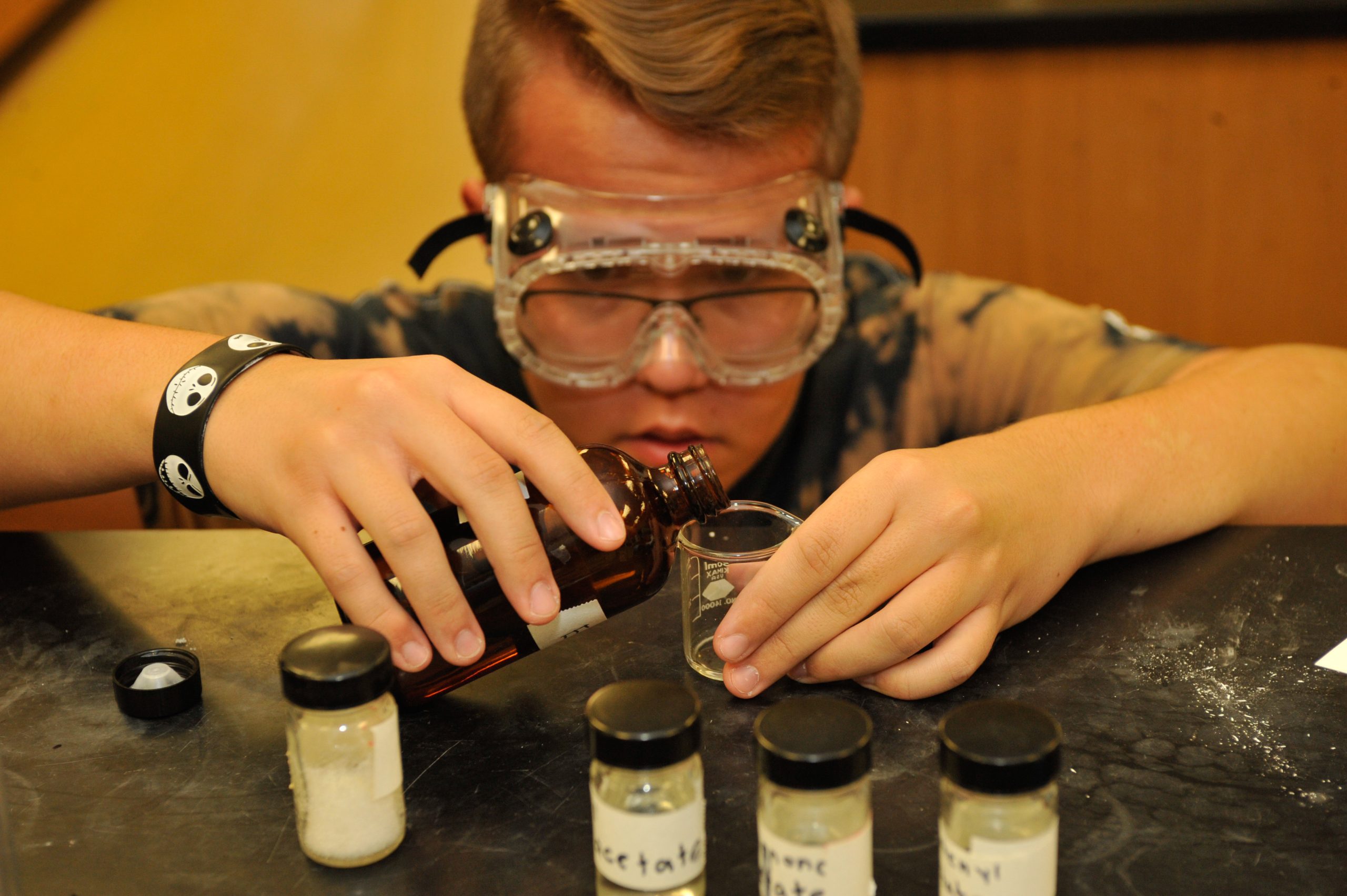 male student with goggles pouring a liquid into small beaker