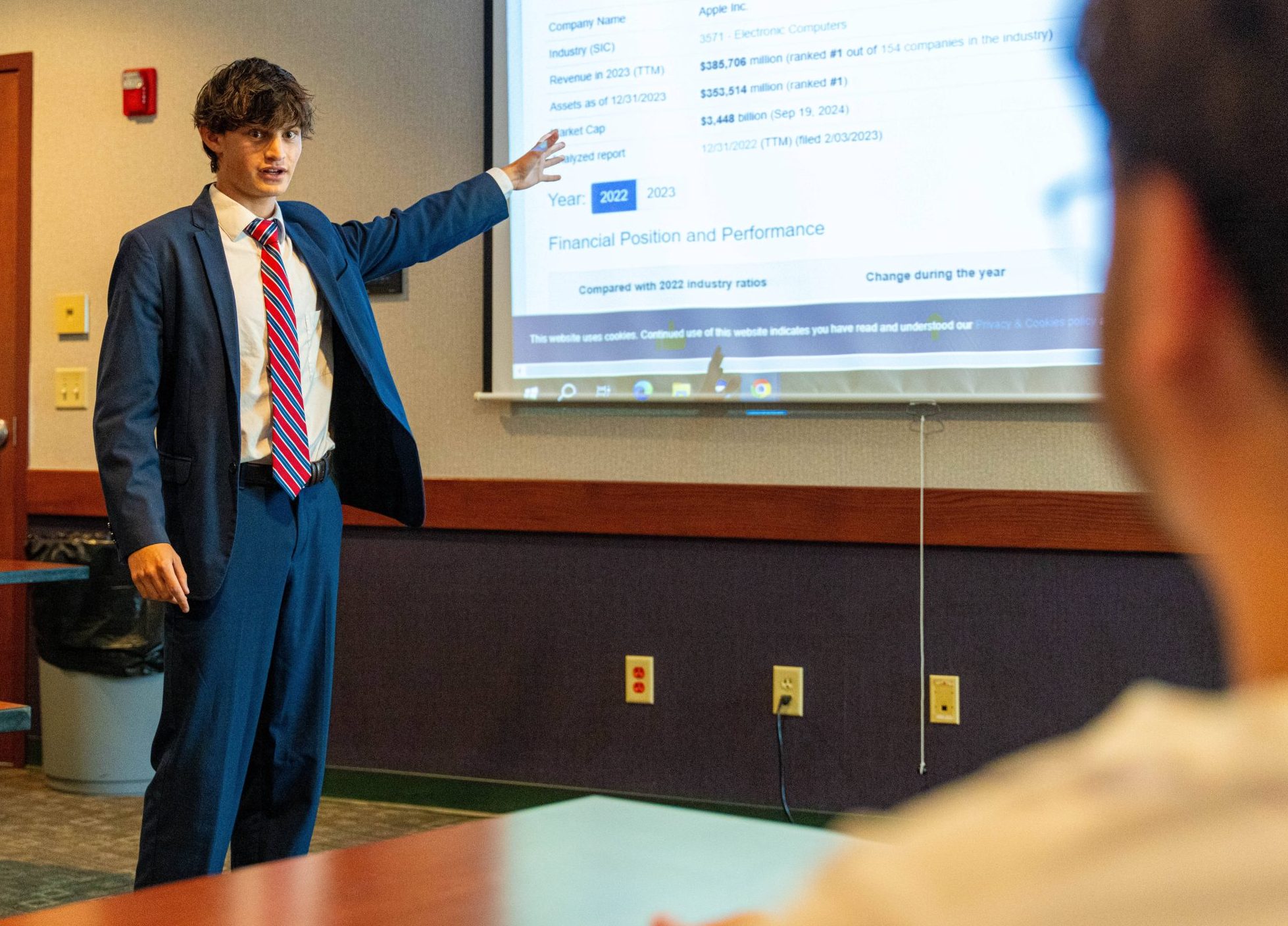 Male student in suit gesturing toward image on a screen for a degree in data analytics.